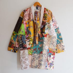 May include: A patchwork kimono-style jacket with a white collar and tie closure. The jacket features a variety of colorful fabric patches with floral and geometric patterns. The sleeves are long, and the jacket hangs on a wooden hanger.