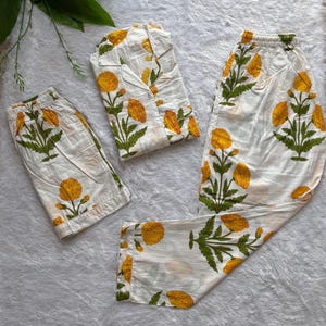 May include: A three-piece pyjama set featuring a button-down shirt, a pair of shorts, and a pair of long trousers. Each piece is made of white fabric with a repeating pattern of yellow flowers and green leaves throughout the entire garment set.