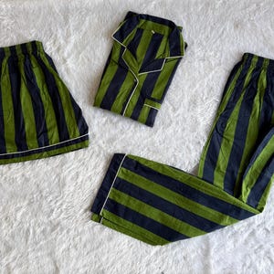 May include: A pajama set consisting of a long-sleeve button-down shirt, long pants, and shorts, all featuring a vertical dark navy blue and olive green striped pattern with white piping trim along the edges, collars, and cuffs of the garments.