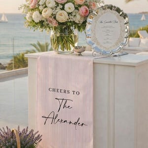 May include: A floral arrangement of white and pink flowers in a clear glass vase sits atop a white bar. A pink and white striped runner with the text "Cheers to The Alexander" drapes over the bar. A silver sign and a basket of lavender are also present.