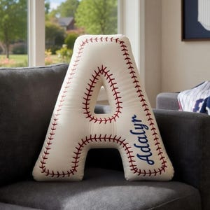 May include: A baseball-themed letter "A" pillow in cream with red stitching, resembling a baseball. The name "Adalyn" is embroidered in blue. The pillow is on a gray couch, with a baseball jersey print in the background.
