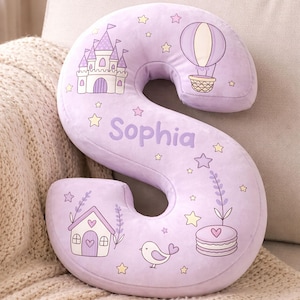 May include: A lavender-colored, plush letter "S" pillow with a whimsical design. It features a castle, hot air balloon, house, bird, and stars. The name "Sophia" is printed on the pillow. This decorative pillow is perfect for a child's room.