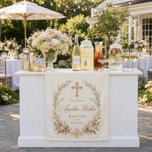 May include: A white rectangular table runner featuring a printed gold cross, floral wreath, and the text God Bless Amelia Parker Baptized 8.25.2026. The runner is draped over a white bar displaying a flower arrangement, glass beverage dispensers, and glasses.
