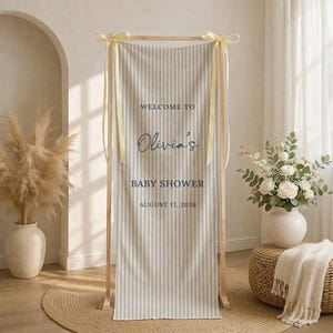 May include: A baby shower welcome sign with vertical gray and white stripes. The sign reads "Welcome to Olivia's Baby Shower August 17, 2026" in navy blue script and block letters. The sign is held by a wooden frame and tied with yellow ribbons.