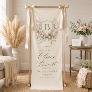 May include: A vertical cream-colored fabric banner hanging from a wooden stand with beige ribbons. It features a floral wreath surrounding the letter B, with the text Welcome to Olivia Bennett's Bridal Shower, Saturday, May 24, 2026.