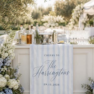 May include: A light blue and white striped fabric runner draped over a bar, featuring the dark blue printed text "CHEERS TO The Harringtons 09.15.2026". The runner is positioned in front of a collection of glass liquor bottles, a metal ice bucket, and clear glasses.