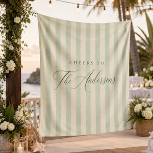 May include: A large fabric backdrop featuring vertical light green and white stripes. The text "CHEERS TO" is printed in a simple sans-serif font above the cursive script text "The Andersons" centered on the fabric surface.