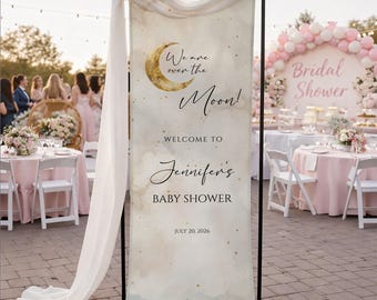 May include: A baby shower banner with the text "We are over the Moon!" and "Welcome to Jennifer's Baby Shower" with the date July 20, 2026. The banner has a gold crescent moon graphic and is set up on a black metal stand.