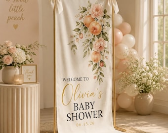May include: A cream-colored baby shower banner with a gold frame, featuring peach and floral illustrations. The banner reads "Welcome to Olivia's Baby Shower" with the date 06.15.26. Cream bows adorn the top corners.