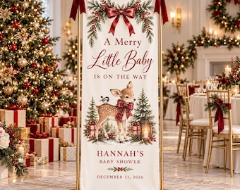 May include: A vertical cream-coloured fabric banner suspended on a gold metal stand with red bows. It features a fawn, pine trees, gifts, and a lantern. Text reads: A Merry Little Baby IS ON THE WAY, HANNAH'S BABY SHOWER, DECEMBER 13, 2026.