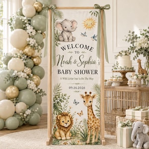 May include: A vertical fabric banner on a wooden stand featuring a baby elephant, lion, and giraffe illustration with the text Welcome to Noah & Sophia's Baby Shower, A Wild Little One Is On The Way, and the date 09.14.2026, decorated with green ribbons.
