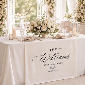 May include: A white rectangular table runner featuring the black printed text THE Williams, STELLA & JAMES, and the date 08.09.2026. The runner is draped over a white tablecloth set with a floral centerpiece, candles, and place settings.