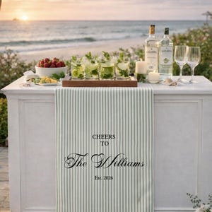 May include: A beachside bar setup with cocktails, fruit, and liquor bottles. The bar features a striped runner with the text "Cheers to The Williams Est. 2026." String lights hang above the ocean at sunset.