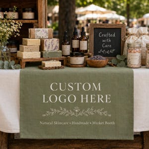 May include: A sage green table runner featuring the white text CUSTOM LOGO HERE with a floral line art graphic and the words Natural Skincare, Handmade, Market Booth, displayed on a table with assorted skincare products and a chalkboard sign.