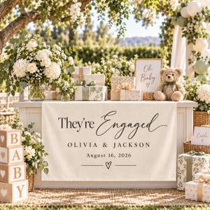 May include: A cream-colored fabric banner featuring the black text "They're Engaged," "OLIVIA & JACKSON," and "August 16, 2026" with a small heart icon. The banner is displayed on a table decorated with white floral arrangements, gift boxes, and a teddy bear.