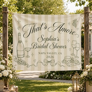 May include: A rectangular banner with a beige and white vertical striped pattern featuring the text That's Amore Sophia's Bridal Shower NAPA VALLEY, CA 2026. The design includes illustrations of a champagne bottle, wine glasses, grapes, and a cheese board.