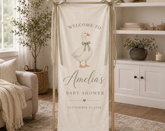May include: A vertical cream-colored fabric banner suspended from a wooden stand with sage green ribbons. The banner features a white goose illustration, the text "WELCOME TO Amelia's BABY SHOWER" and "SEPTEMBER 21, 2026" in sage green.