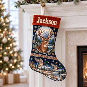 May include: A dark blue Christmas stocking with a rust-colored velvet cuff featuring the name Jackson in white text. The stocking displays a central oval portrait of a deer and a lower scene of two mallard ducks flying over a snowy river.