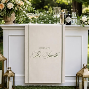 May include: A cream-colored fabric table runner draped over a white bar, featuring dark green vertical border lines and the text "CHEERS TO The Smith" printed in a dark green serif and script font, centered on the front panel of the runner.