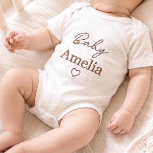 Könnte beinhalten: Ein weißer kurzärmeliger Babybody mit braunem Sticktext, der Baby Amelia über einem kleinen Herzumriss zeigt. Das Kleidungsstück hat einen Rundhalsausschnitt und Druckknöpfe im Schrittbereich, gestaltet für Säuglinge mit einem schlichten Design.