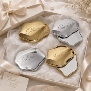 May include: Four irregular, cloud-shaped compact mirrors with metallic finishes in gold and silver. Each mirror is engraved with a name: Olivia, Charlotte, Sophia, or Evelyn. The mirrors are arranged inside a gift box with white tissue paper.