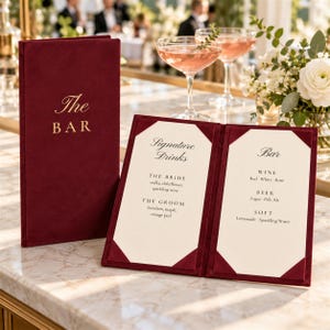 May include: A deep burgundy velvet menu cover with gold embossed text reading The BAR, alongside an open matching menu holder displaying Signature Drinks and Bar selections including wine, beer, and soft drinks on cream-colored cardstock inserts.