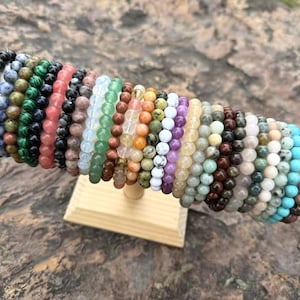 Puede incluir: Una exhibición de pulseras de cuentas en varios colores, incluyendo negro, verde, rosa y azul. Las pulseras están hechas de diferentes tipos de piedras y están dispuestas en un soporte de madera.