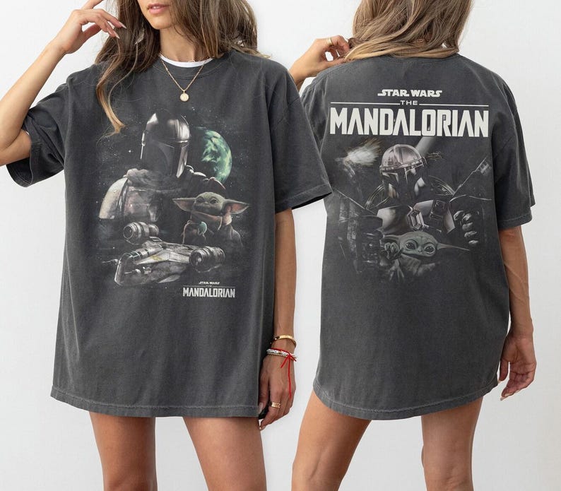 May include: Dark gray t-shirt featuring a graphic from The Mandalorian. The front design includes the Mandalorian, Grogu, and a spaceship. The back of the shirt displays the Star Wars logo and the show's title. The shirt is a relaxed fit.