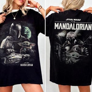 May include: Black t-shirt featuring a graphic of The Mandalorian and Grogu from Star Wars. The front design shows the characters and a spaceship against a space background. The back of the shirt displays the Star Wars logo and "The Mandalorian".
