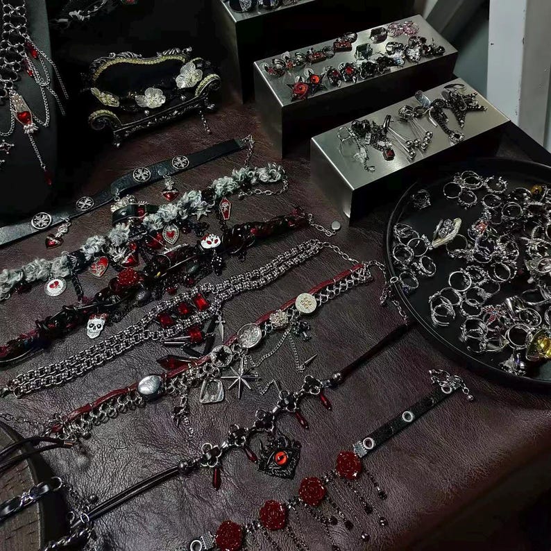 May include: A collection of gothic-style jewelry, including chokers, rings, and bracelets, displayed on a dark brown surface. The chokers feature red and silver accents, skulls, and charms. Rings are displayed in a tray and on metal display boxes. A small black couch is in the background.