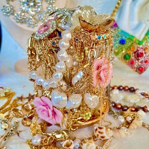 Puede incluir: Una colección de joyas doradas, que incluye collares, pulseras y pendientes, que se desborda de un recipiente de vidrio decorativo. Las joyas presentan perlas, conchas marinas y acentos de rosas rosas. En el fondo se ve un collar de pedrería brillante.