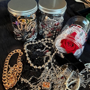 May include: An assortment of jewelry, including necklaces, bracelets, and rings, is displayed. Two jars filled with various pieces of jewelry are visible, along with a jar containing a red rose. The jewelry includes gold, silver, black, and red pieces.