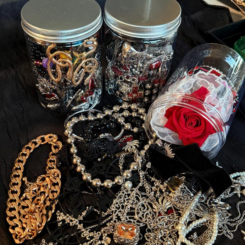 May include: An assortment of jewelry, including necklaces, bracelets, and rings, is displayed. Two jars filled with various pieces of jewelry are visible, along with a jar containing a red rose. The jewelry includes gold, silver, black, and red pieces.