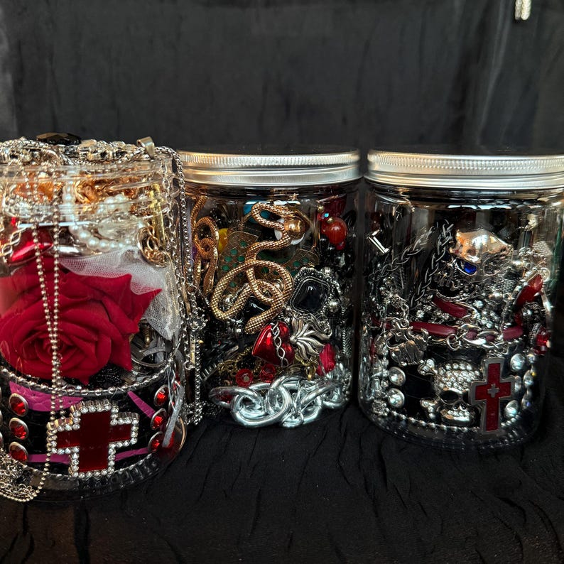 May include: Three glass jars filled with various jewelry pieces. The jars contain necklaces, bracelets, and other accessories in silver, gold, and red tones. One jar features a red rose and a cross design.