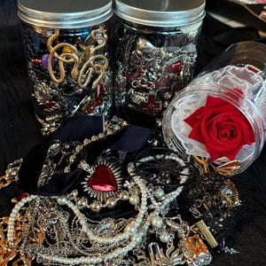 May include: An assortment of jewelry, including necklaces, earrings, and brooches, spills from glass jars and a black velvet surface. A red rose rests in a clear container. The jewelry features silver, gold, and pearl accents, with some pieces incorporating red gemstones.