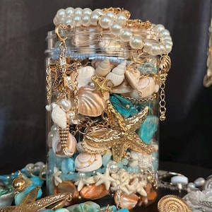Könnte beinhalten: Ein klares Glasgefäß gefüllt mit Muscheln, goldenen Seesternen, Perlen und türkisfarbenen Steinen. Goldfarbener Schmuck, darunter Armbänder und Charms, verziert das Glas. Das Glas ist überfüllt mit dekorativen Strandartikeln.