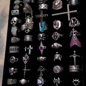 May include: A collection of silver rings with various designs, including skulls, crosses, and geometric shapes, displayed on a black surface. Some rings feature gemstones and intricate detailing, showcasing a gothic or alternative aesthetic. The rings vary in size and style.
