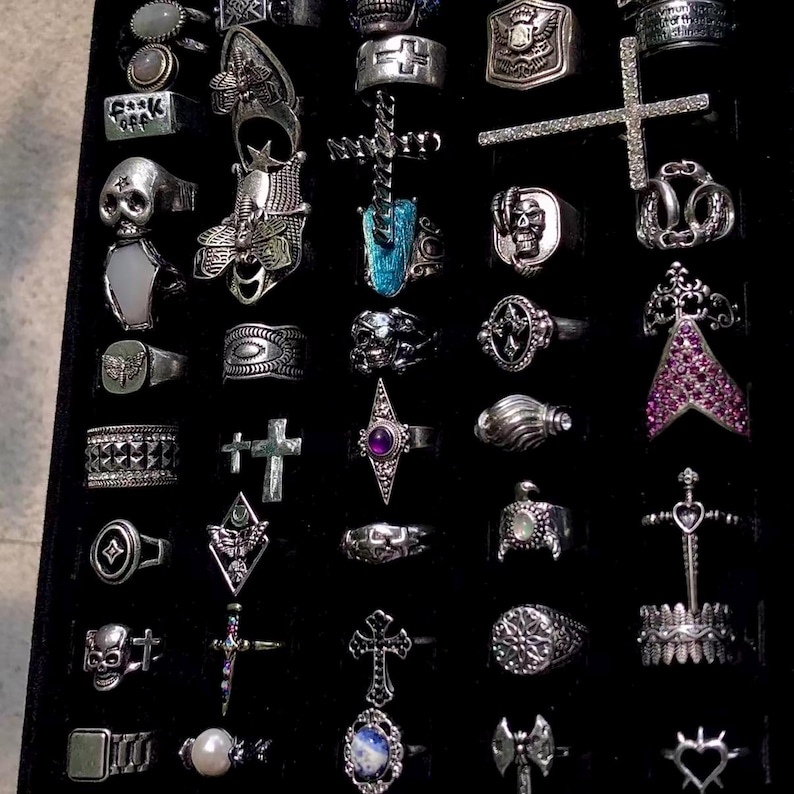 May include: A collection of silver rings with various designs, including skulls, crosses, and geometric shapes, displayed on a black surface. Some rings feature gemstones and intricate detailing, showcasing a gothic or alternative aesthetic. The rings vary in size and style.