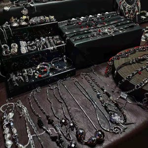 May include: A collection of silver-toned necklaces, bracelets, and earrings displayed on a dark brown surface. The jewelry includes various designs, such as chains, beads, and pendants. Some pieces are arranged on black display boxes, showcasing a variety of styles and embellishments.