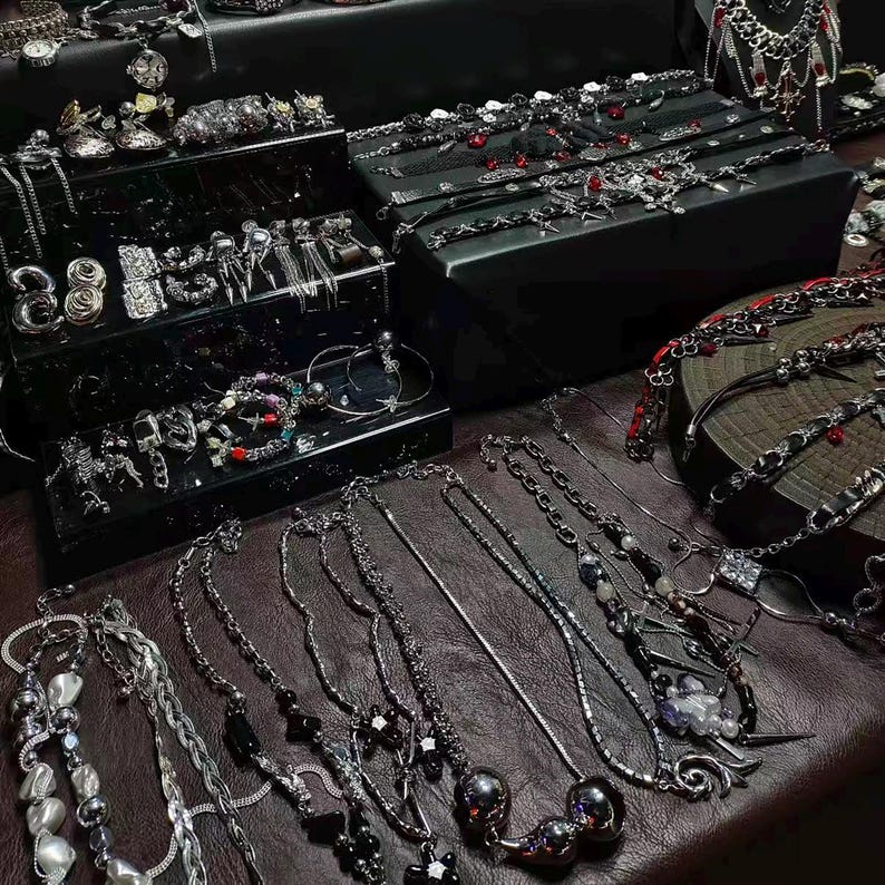 May include: A collection of silver-toned necklaces, bracelets, and earrings displayed on a dark brown surface. The jewelry includes various designs, such as chains, beads, and pendants. Some pieces are arranged on black display boxes, showcasing a variety of styles and embellishments.