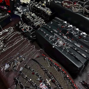 May include: An assortment of gothic-style jewelry, including necklaces, bracelets, and chokers, displayed on a dark wooden surface and black boxes. The jewelry features silver-toned metal, black leather, and red accents. Various rings and other accessories are also visible.