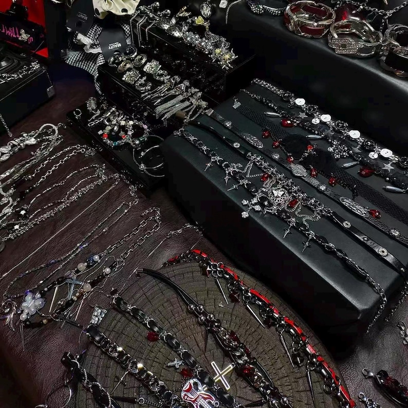 May include: An assortment of gothic-style jewelry, including necklaces, bracelets, and chokers, displayed on a dark wooden surface and black boxes. The jewelry features silver-toned metal, black leather, and red accents. Various rings and other accessories are also visible.