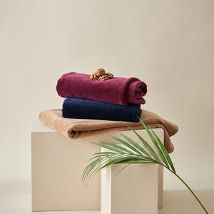 May include: A stack of three folded blankets in deep burgundy, navy blue, and tan colors. The top burgundy blanket is topped with a small cluster of walnuts. Two orange slices are placed near the base of the display, and a green palm leaf.