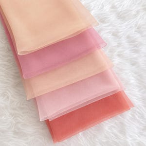 Peut inclure: Une pile de cinq échantillons de tissu rectangulaires en matière transparente et diaphane. Les couches sont disposées en dégradé de couleurs, allant d'un pêche pâle et rose doux à un rouge corail plus profond et saturé en bas de la pile.