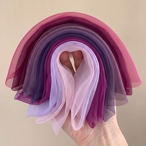 May include: A collection of layered sheer fabric squares arranged in a semi-circular fan shape. The fabric pieces transition in color from a deep magenta and dark purple on the outer edges to lighter shades of lavender and pale pink in the center.