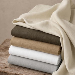 Puede incluir: Una pila de cinco piezas de tela dobladas en crema, verde oliva oscuro, bronceado, blanco y gris claro. La tela crema superior está drapeada sobre el lado de la pila, revelando una textura de tejido de sarga diagonal visible en todos los textiles.