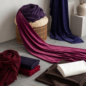 May include: A collection of velvet fabric pieces in various colors including deep purple, magenta, dark blue, burgundy, brown, and off-white. The fabrics are displayed as draped lengths, folded stacks, and rolled bundles, showcasing the soft texture.