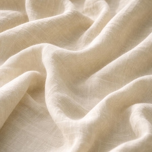 May include: Close-up of a cream-colored fabric with a soft, flowing texture. The fabric is draped in gentle folds, creating a sense of movement and depth. The weave is visible, suggesting a natural fiber textile.