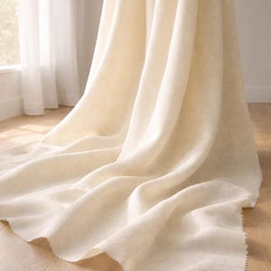 May include: Cream-colored linen curtain panel with a textured weave. The fabric drapes in soft folds, catching the light. The curtain is on a light wood floor, with a sheer white curtain in the background.