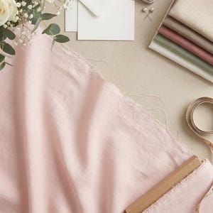 May include: A flat lay featuring a pale pink fabric, white envelopes, and a bouquet of white roses. Other items include a ring box, fabric swatches in various colors, a measuring tape, and a pair of pearl earrings. The scene is set on a neutral background.